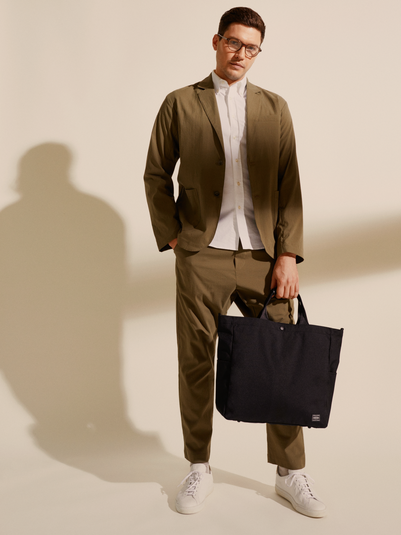 Man wearing olive Smock x Monocle Yoyogi seersucker suit with white shirt and sneakers holding black tote bag