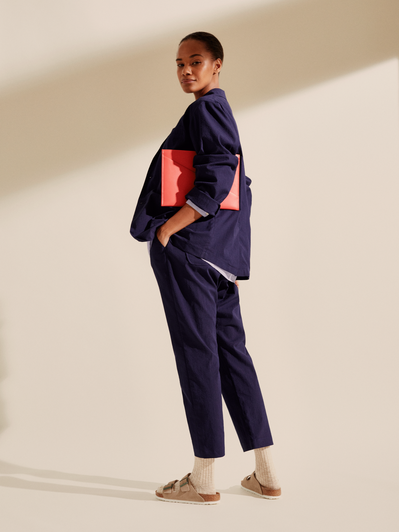 Woman wearing navy Smock x Monocle Yoyogi seersucker suit with coral clutch and Birkenstock sandals