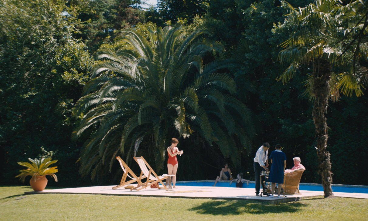 Poolside scene with palm trees, lounge chairs, and people around a swimming pool in a tropical setting