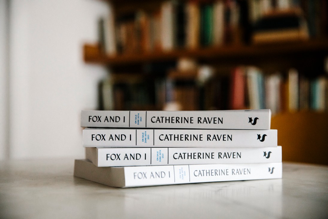 Stack of four copies of 'Fox and I' by Catherine Raven published by Spiegel & Grau, displayed on a surface with bookshelves blurred in background.