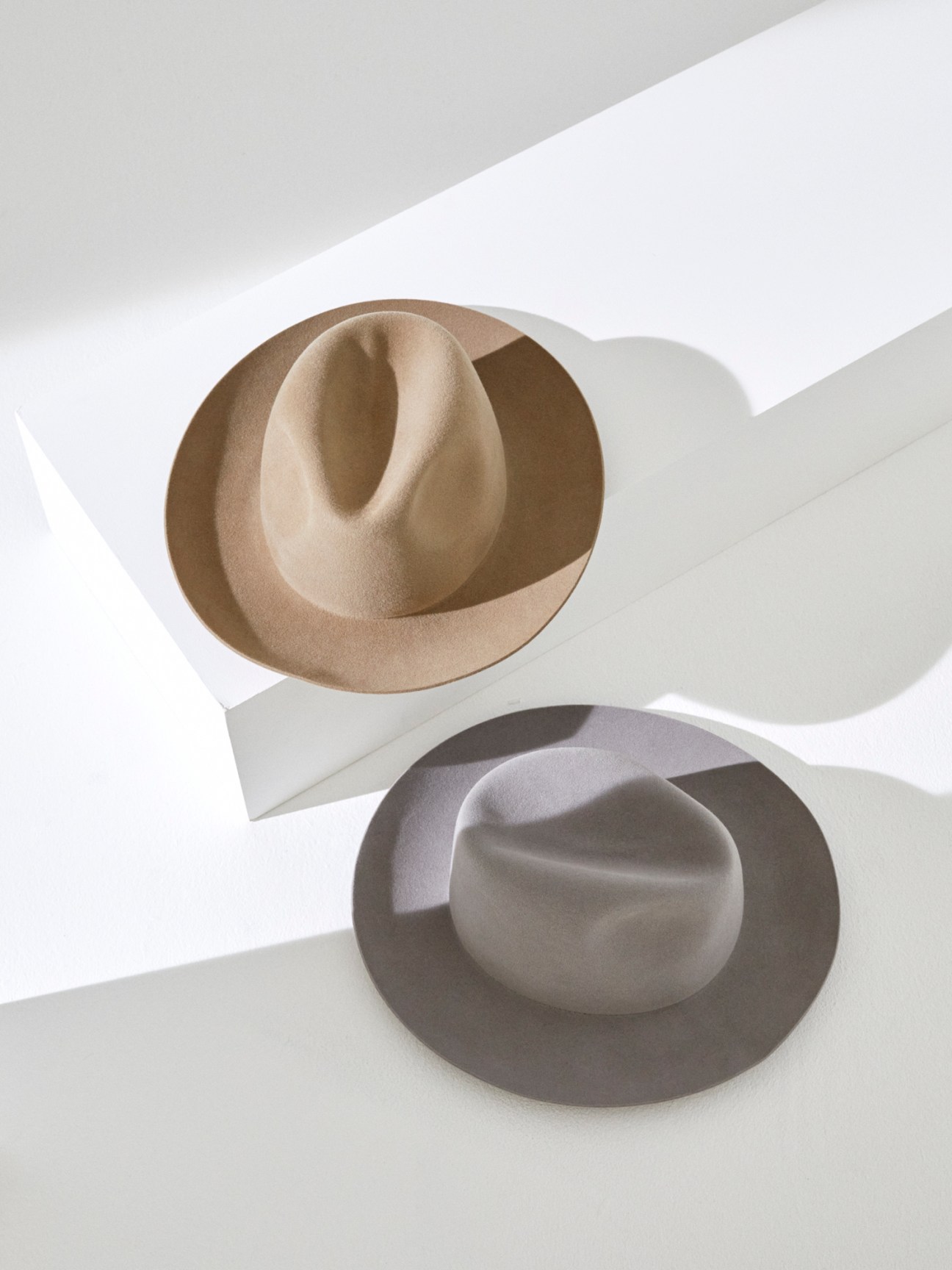 Two felt hats in beige and gray tones photographed against white surfaces with dramatic shadows