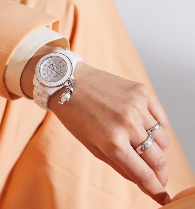 Woman's hand wearing white Chanel J12 watch and Coco Crush rings against peach fabric