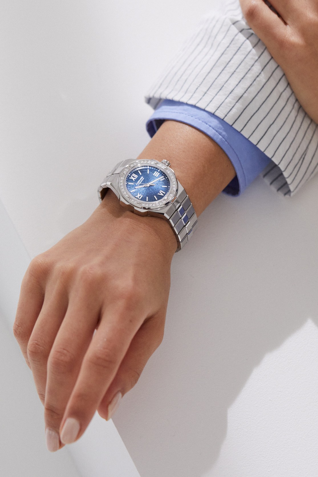 A hand wearing a Chopard Alpine Eagle watch with blue dial and diamond bezel, dressed in a striped shirt and blue jacket