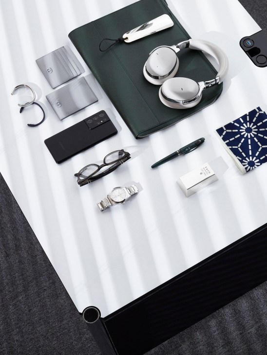 Flat lay of luxury accessories including Montblanc headphones, Dior card cases, Persol glasses, Tudor watch, and other designer items on white background.