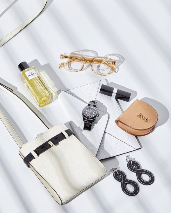Luxury accessories styled flat lay including Chanel fragrances, Prada glasses, Saint Laurent baby satchel, Chanel J12 watch, and Hermès earrings.
