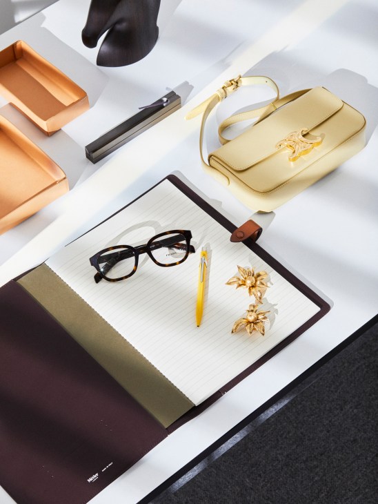 Styled flat-lay of luxury accessories including a gold handbag, notebook, glasses, pen, earrings and incense burner on white surface.