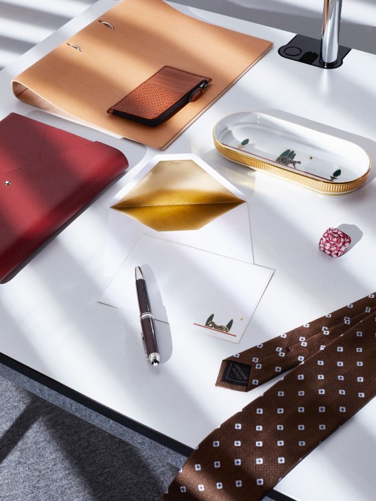 Styled desk arrangement featuring leather accessories, stationery, and luxury items including notebooks, pen, tie, and decorative objects.