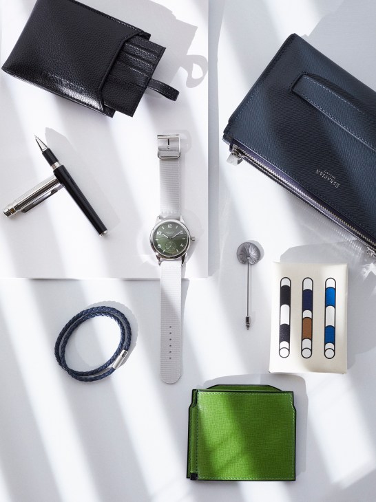 Flat lay of accessories including black leather wallets, a watch with white strap, ballpoint pen, blue bracelet, lapel pin, and colored card holders on white background.