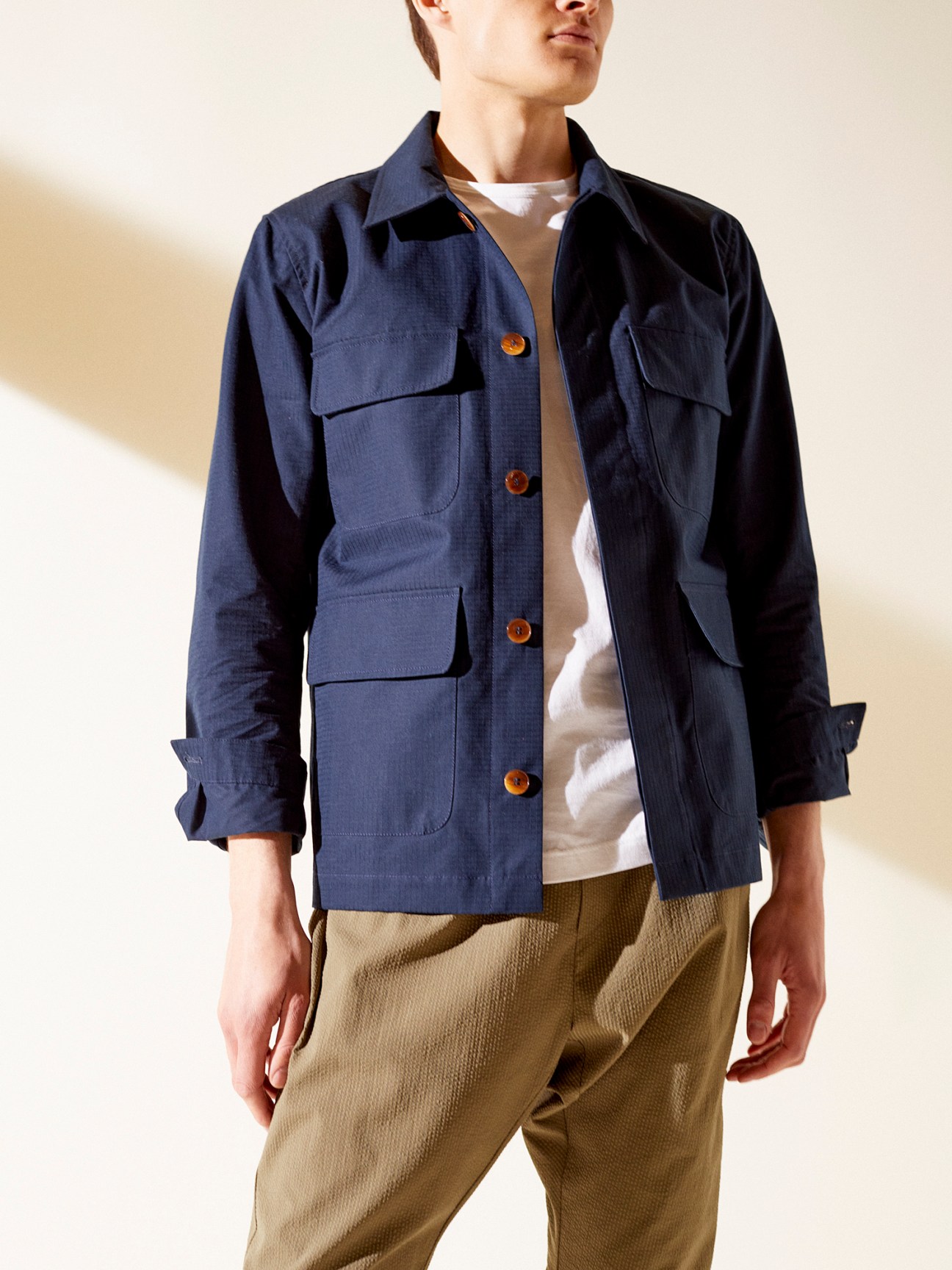 A Kind of Guise x Monocle safari jacket in navy worn over white t-shirt with olive trousers
