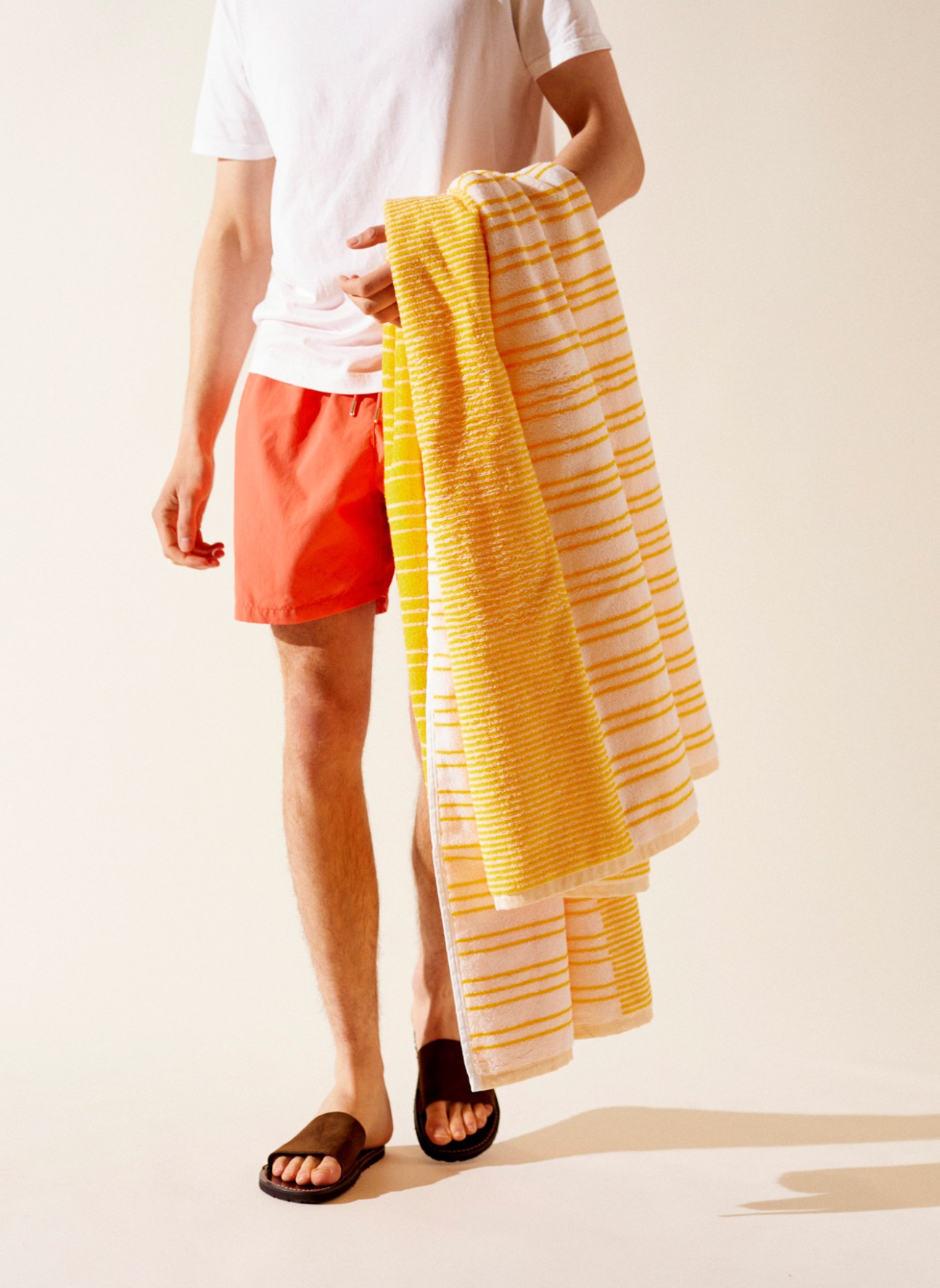 Person holding yellow striped Monocle beach towel wearing coral shorts and black sandals