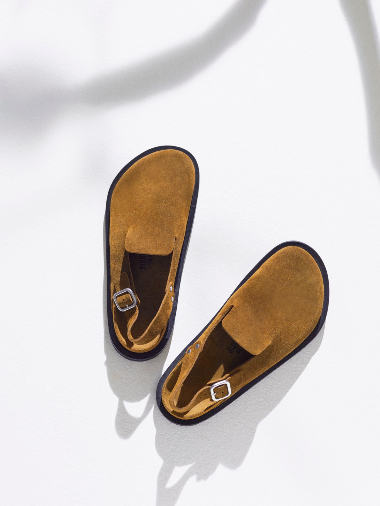 Tan suede sandals with ankle straps and black soles displayed against white background