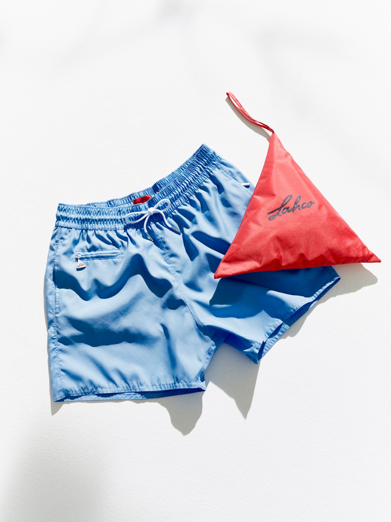 Light blue swim shorts with drawstring waist and coral pink mesh bag laid flat against white background