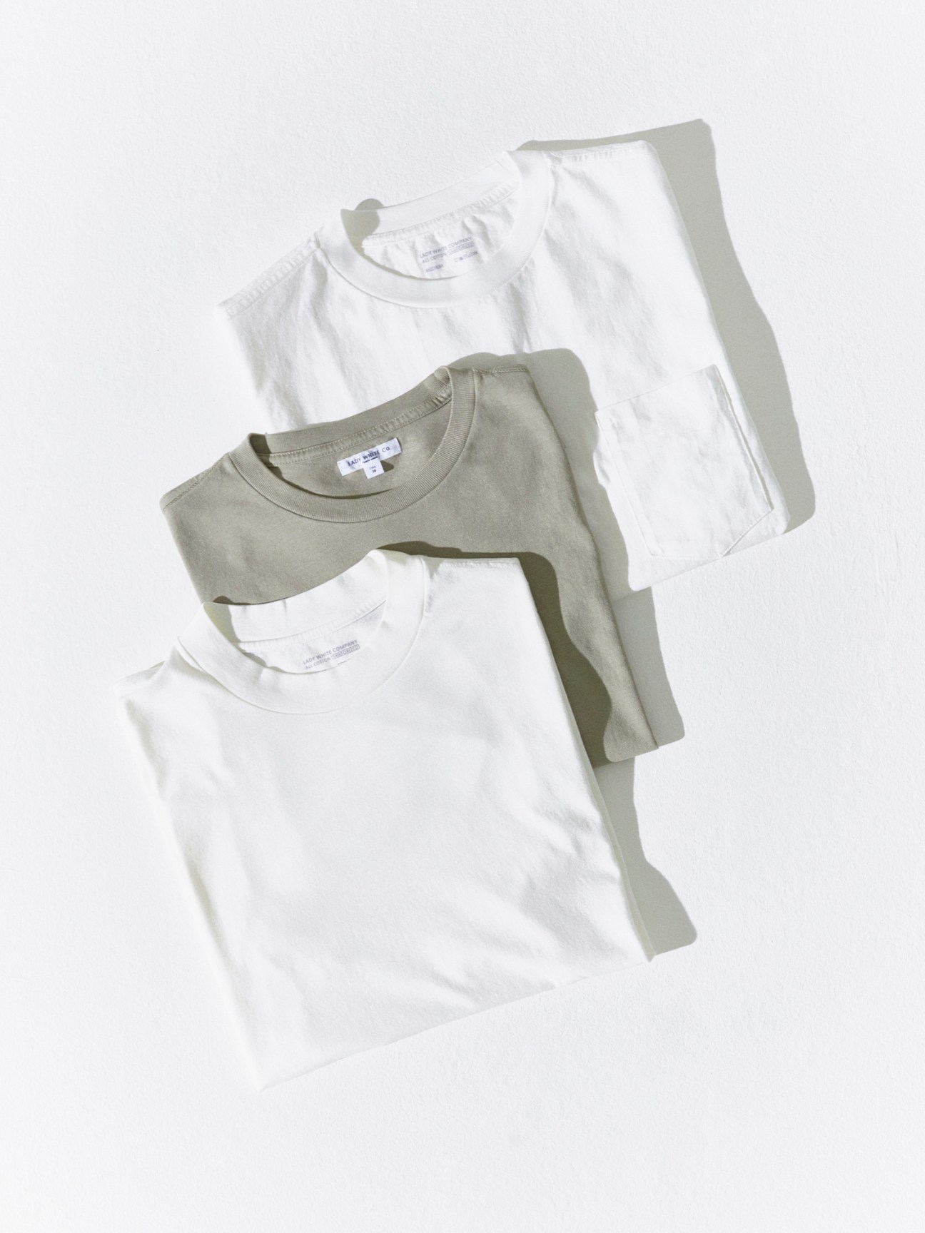 Two white t-shirts and one olive green t-shirt by Lady White Co laid flat against white background
