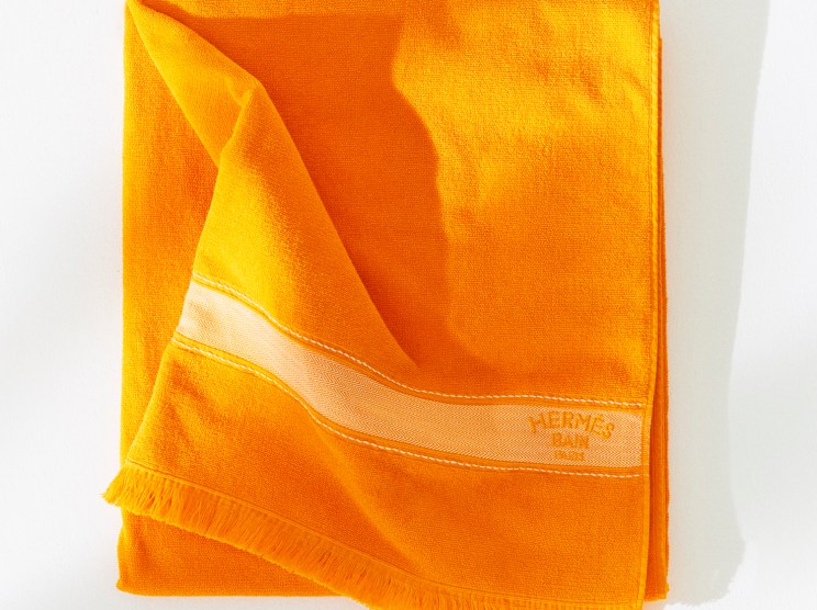 Bright orange Hermès beach towel with fringe edges folded against white background