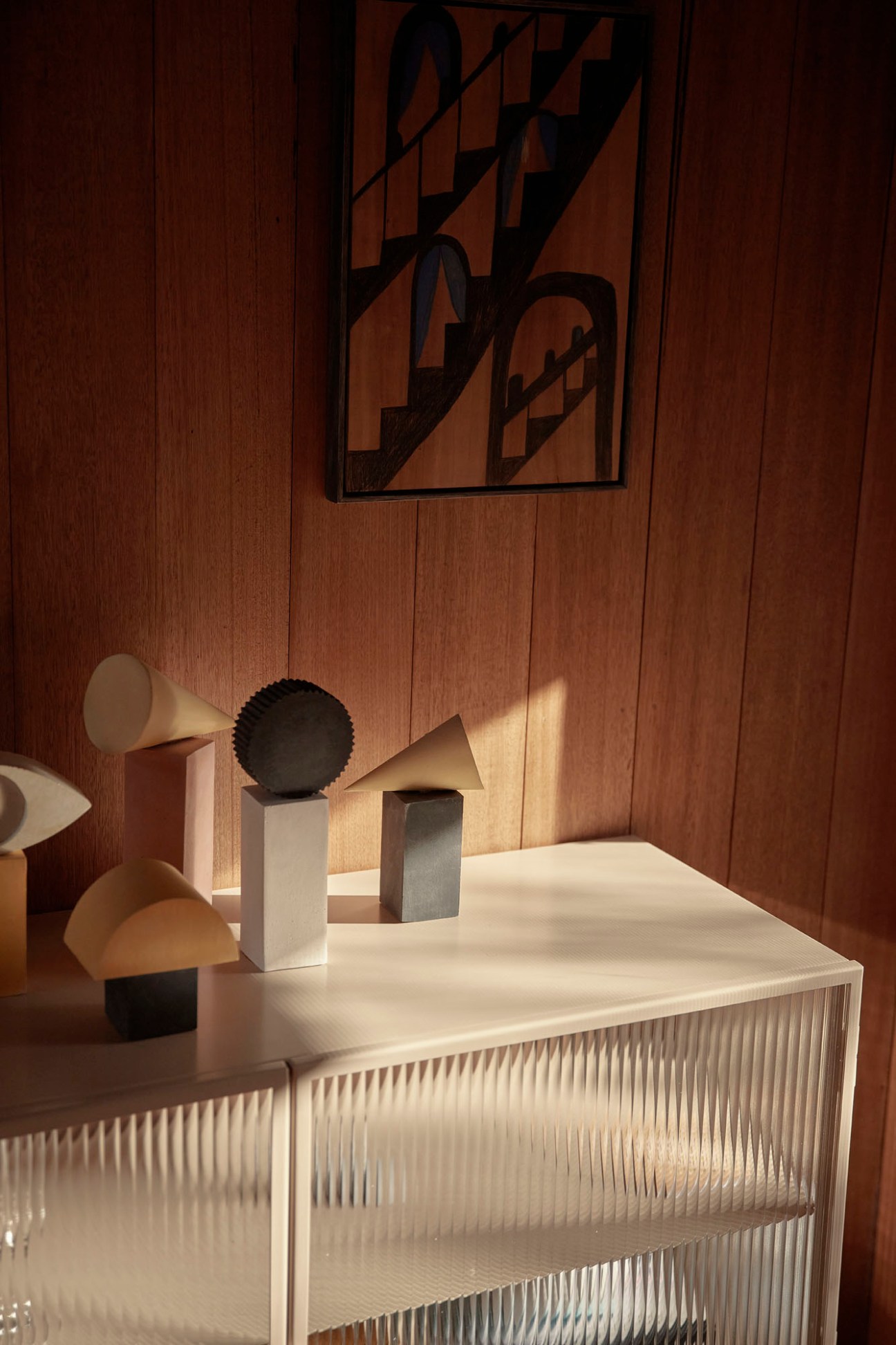 Geometric sculptures by Sean Meilak and framed drawing by Emily Ferretti displayed on a white ribbed cabinet against wooden walls.