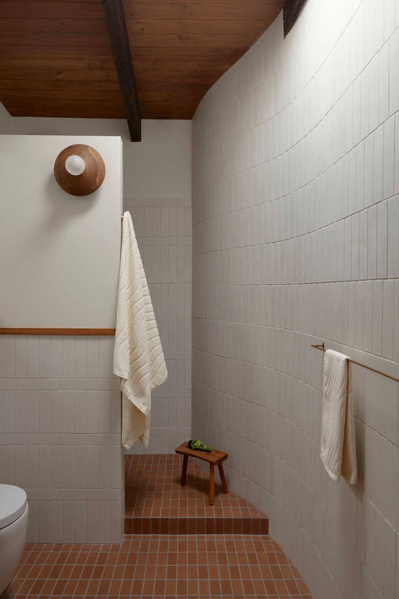 Minimalist en-suite bathroom with white tile walls, terracotta brick flooring, wooden ceiling, and wooden light fixture.