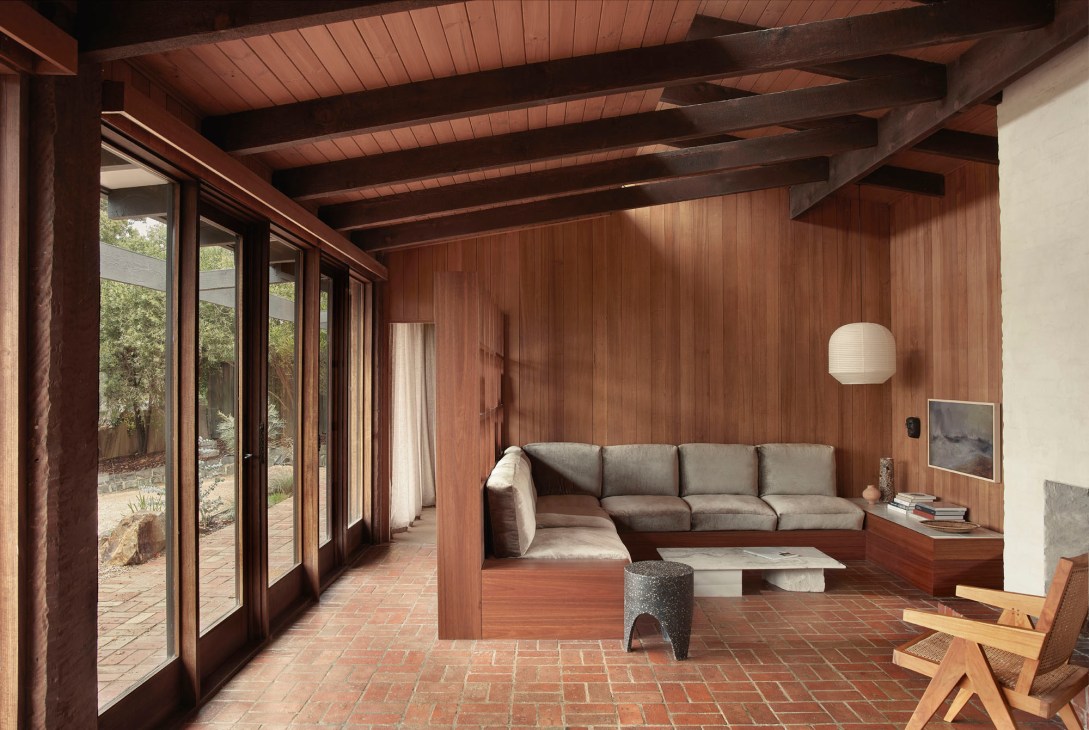 Modernist living room with timber-lined built-in sofa, brick floors, and large glass doors opening to garden.