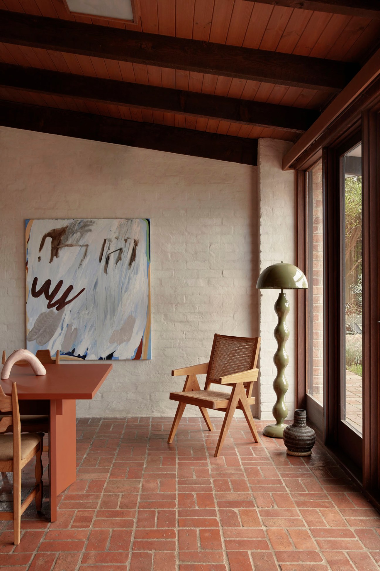 Modernist interior with brick floors, timber ceiling beams, abstract artwork by Elyss McCleary on white wall, mid-century chair and sculptural floor lamp.