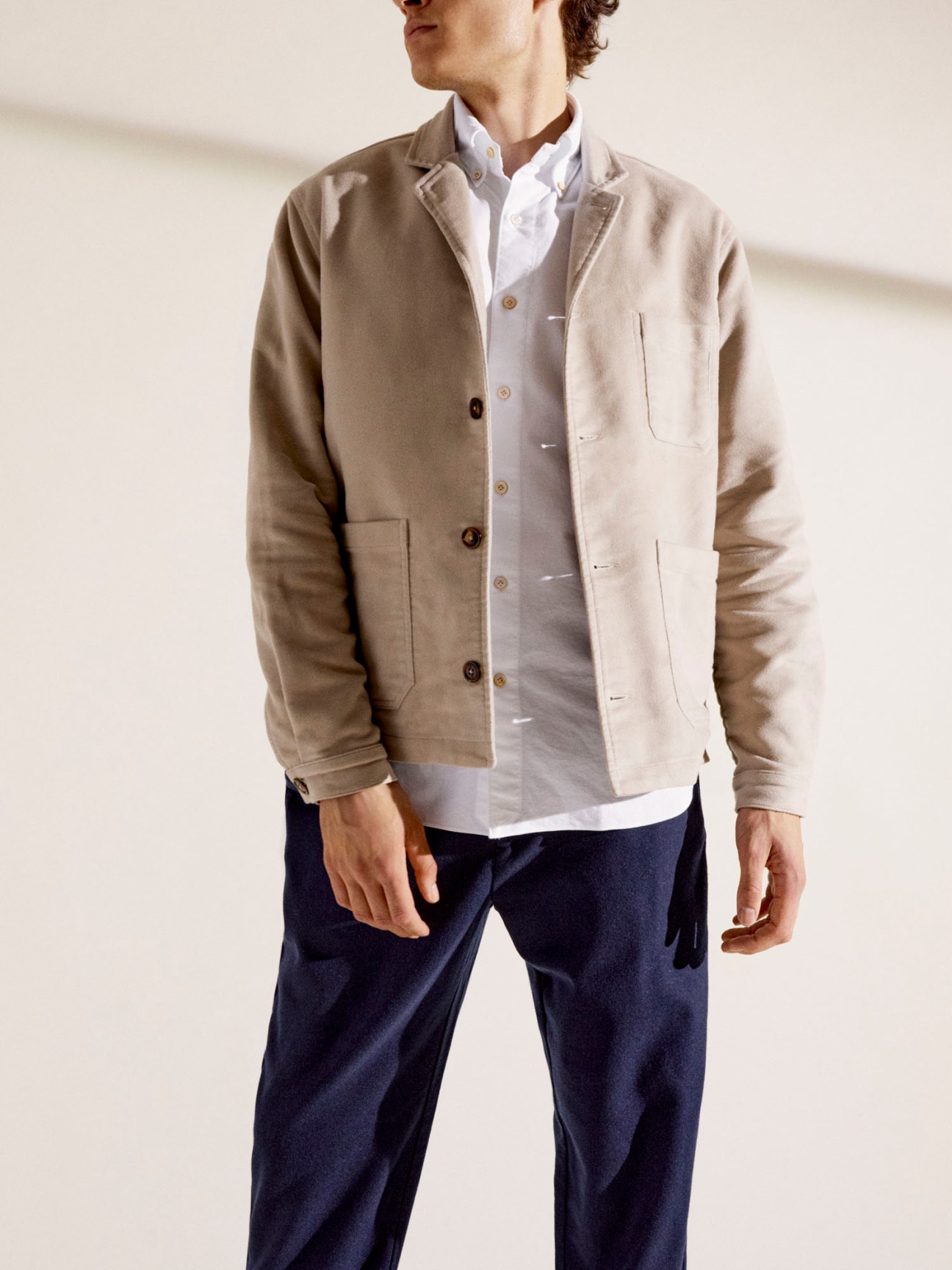 Man wearing beige jacket over white shirt and navy trousers in minimal interior setting