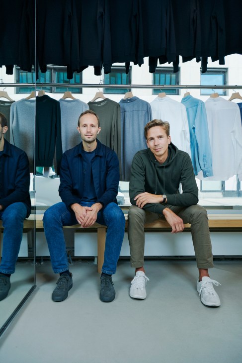 Two men sit on a bench in front of a display of hanging clothing in neutral tones.