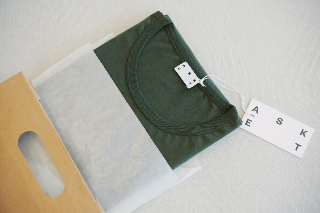 Asket dusty green t-shirt folded with branded label tag on white packaging