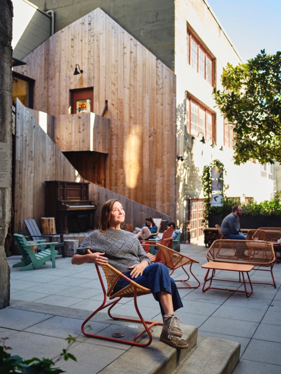 Woman sitting in a modern chair in a sunlit courtyard between renovated buildings with wooden facade.