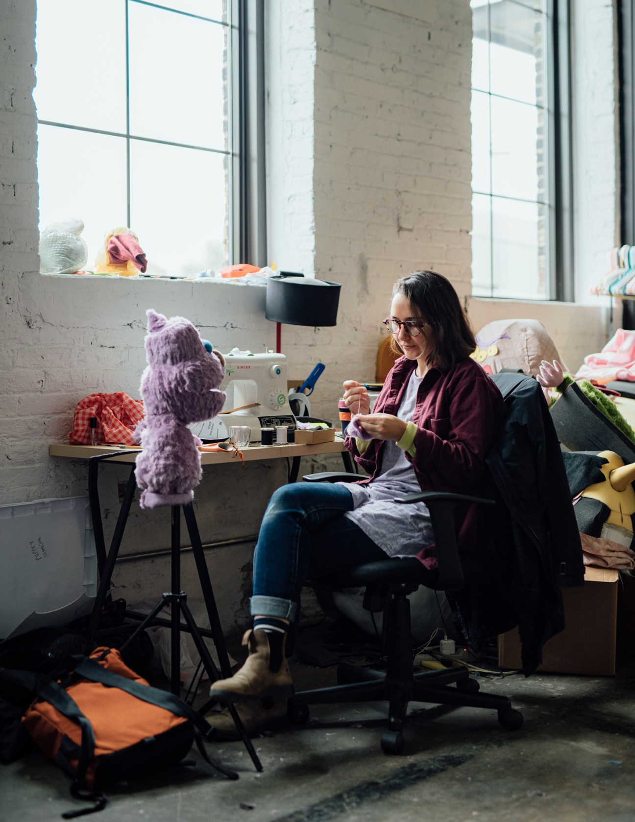 Woman with glasses and burgundy cardigan hand-sewing fabric in bright studio workspace
