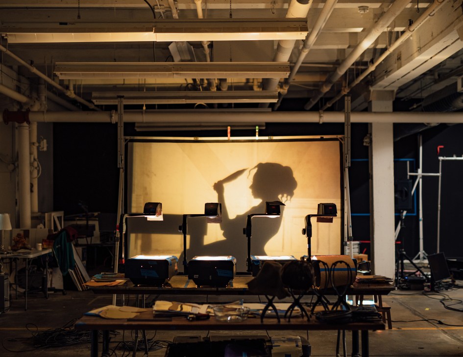 Four overhead projectors casting shadows onto a large screen in a warehouse-like studio space