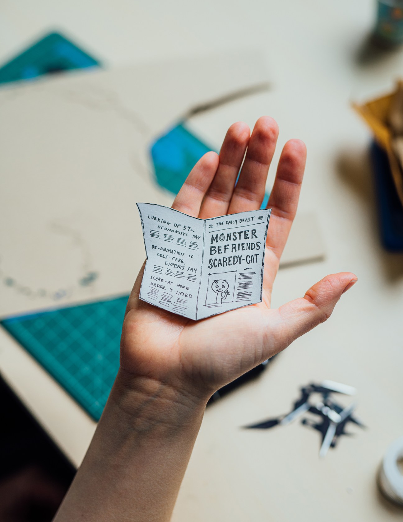 Hand holding a small paper newspaper prop with 'Monster Befriends Scaredy-Cat' headline and simple drawings