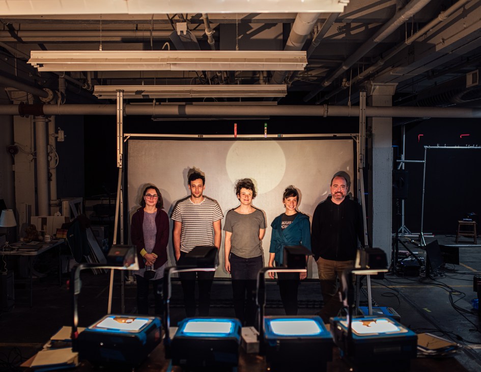 Five Manual Cinema team members standing in their industrial studio workspace with projection equipment
