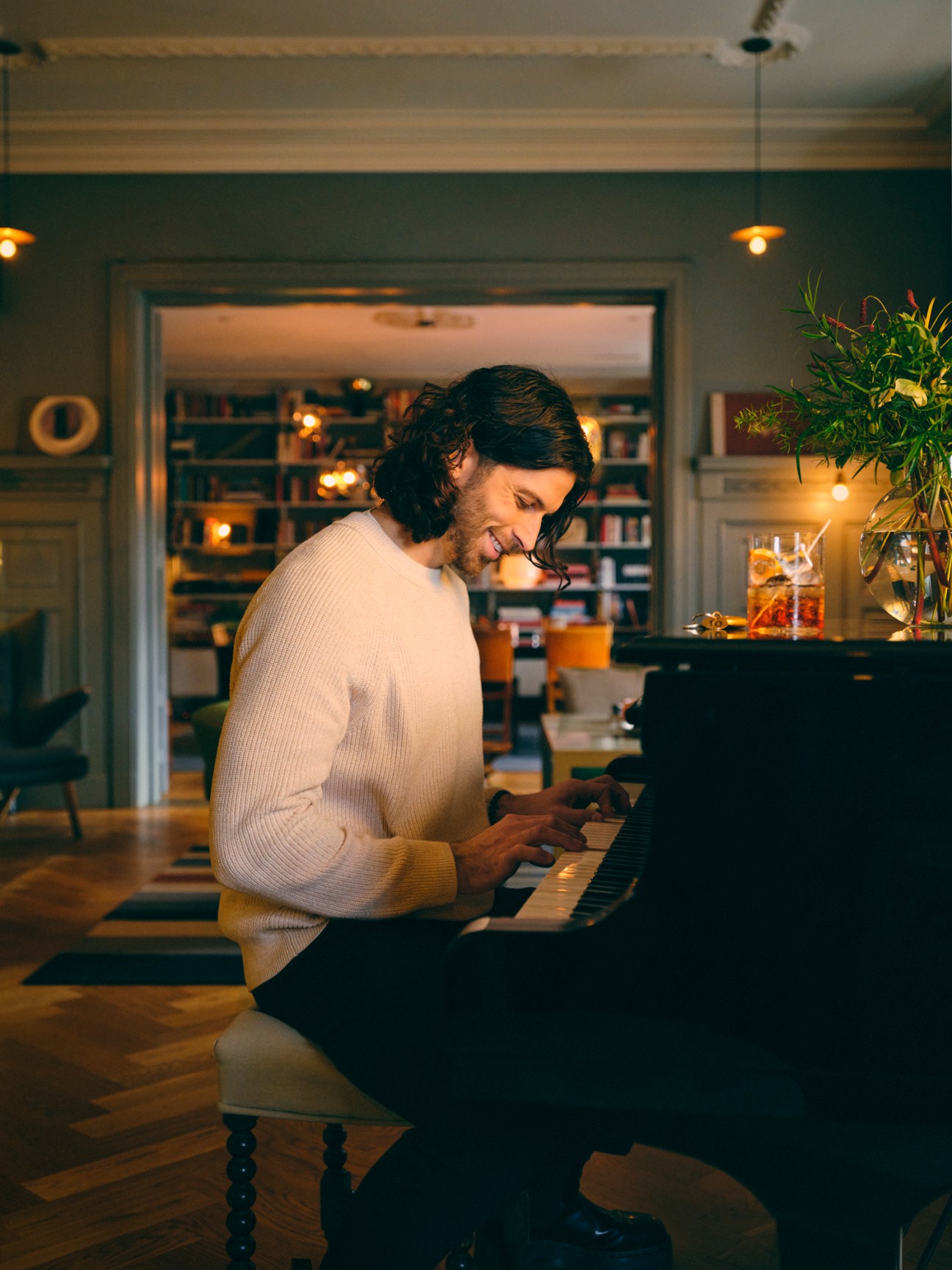 Man in cream sweater playing piano in cozy living room with bookshelves and warm lighting