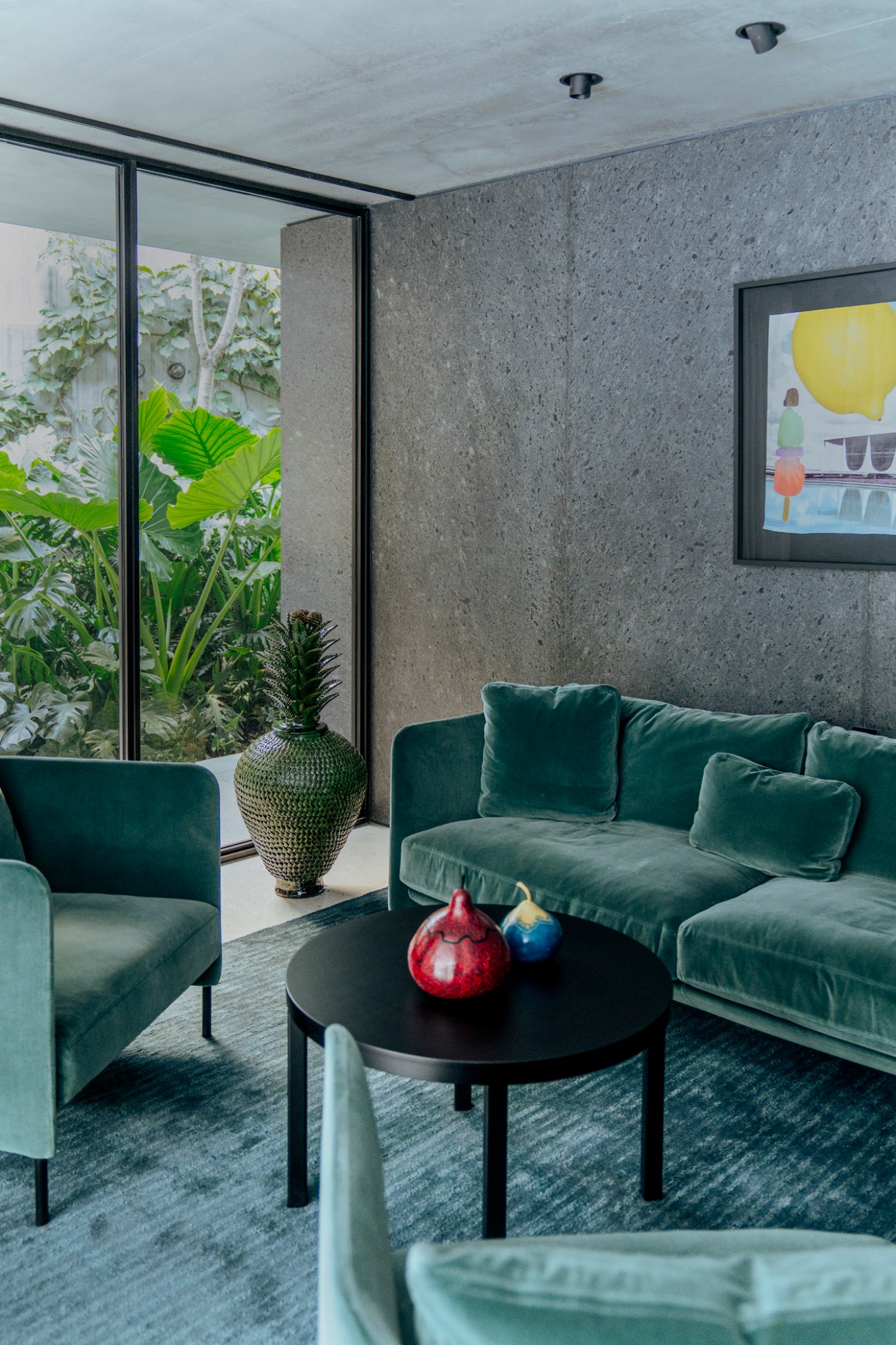 Modern living room with sage green velvet sofa, concrete walls, and large windows overlooking lush garden