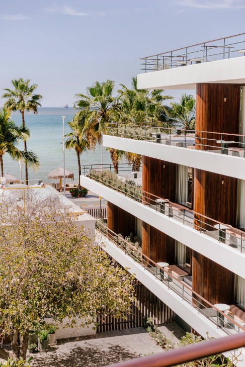 Multi-story beachfront hotel with white balconies, wooden doors, and palm trees overlooking the sea in La Paz.