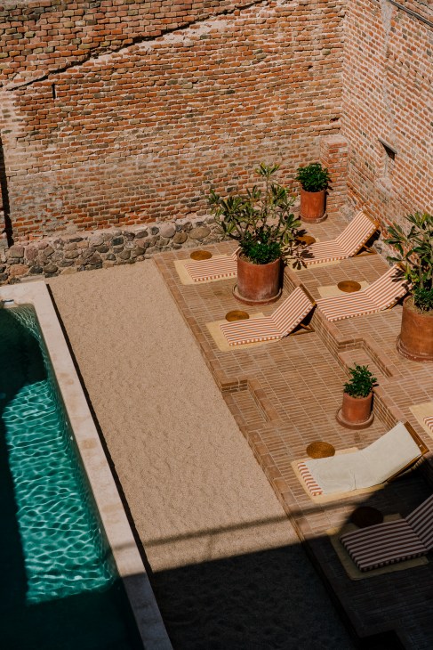 Overhead view of a hotel courtyard with a pool, terracotta planters, striped loungers, and exposed brick walls.