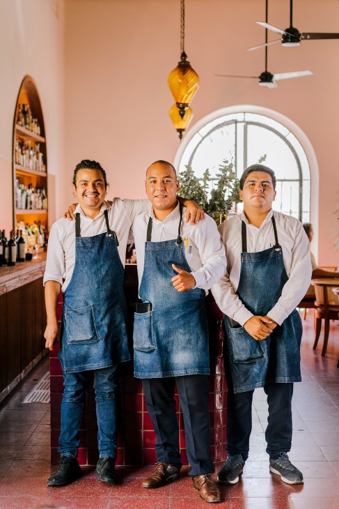 Three male staff members in denim aprons stand smiling in a hotel interior with pink walls and hanging pendant lights.