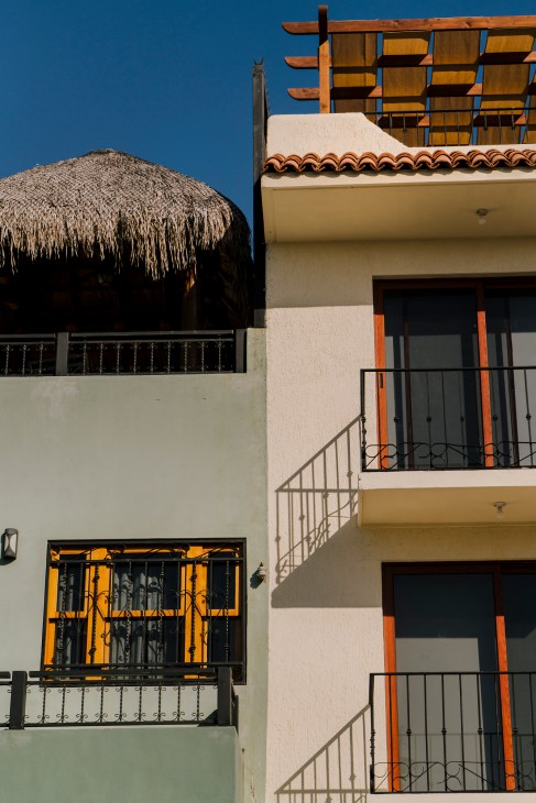 A whitewashed Mexican colonial-style building with orange-trimmed windows and a thatched-roof palapa against a clear blue sky.