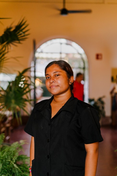 Lupita, a Baja Club hotel janitor, standing in a warm-toned corridor with plants and an arched doorway behind her.