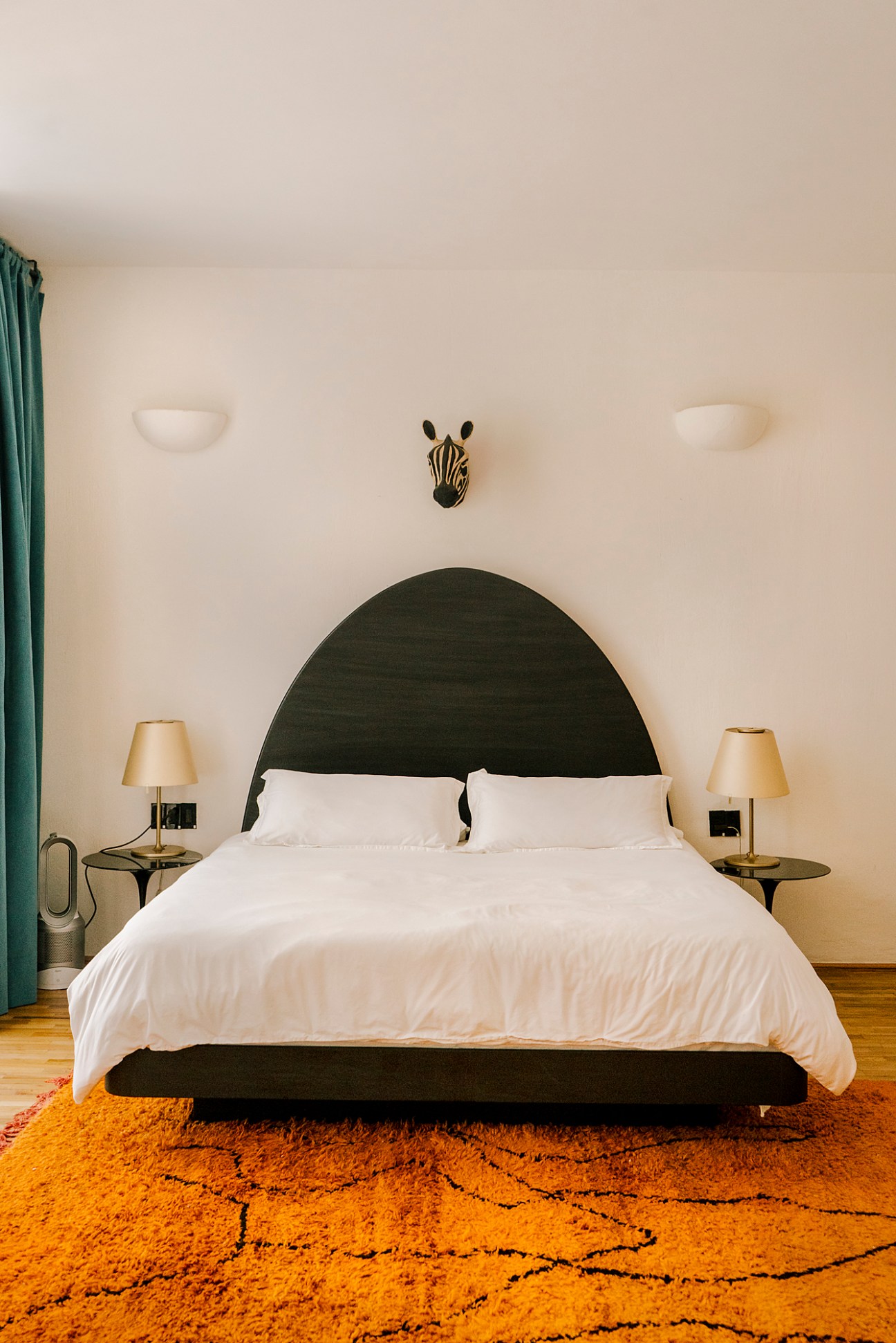 Bedroom with dark green curved headboard, white bedding, brass lamps and orange patterned rug