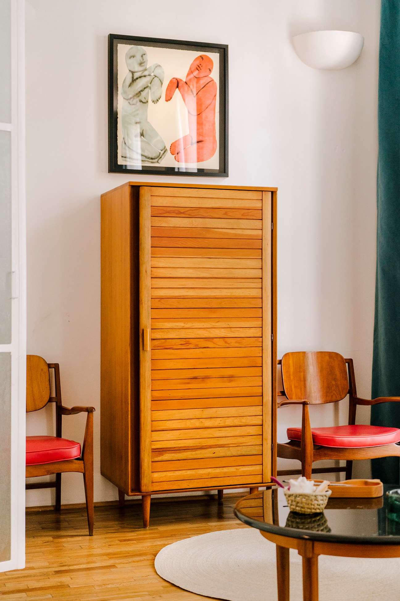 Mid-century modern interior with wooden armoire, red-cushioned chairs, and framed artwork on white wall