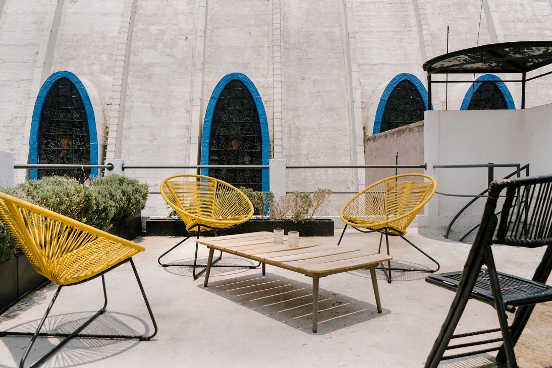 Outdoor terrace with yellow and black woven chairs around wooden table, white building with blue-framed arched windows