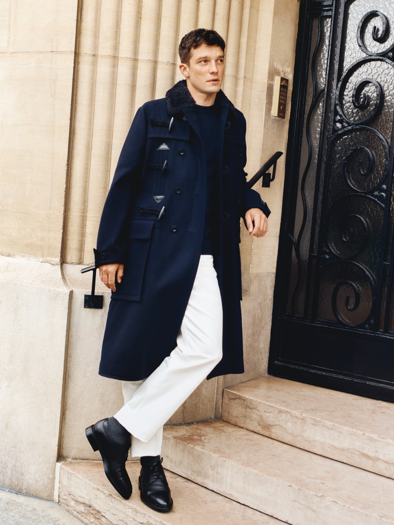 Model in navy coat with black collar over dark jumper with white trousers and black shoes on stone steps