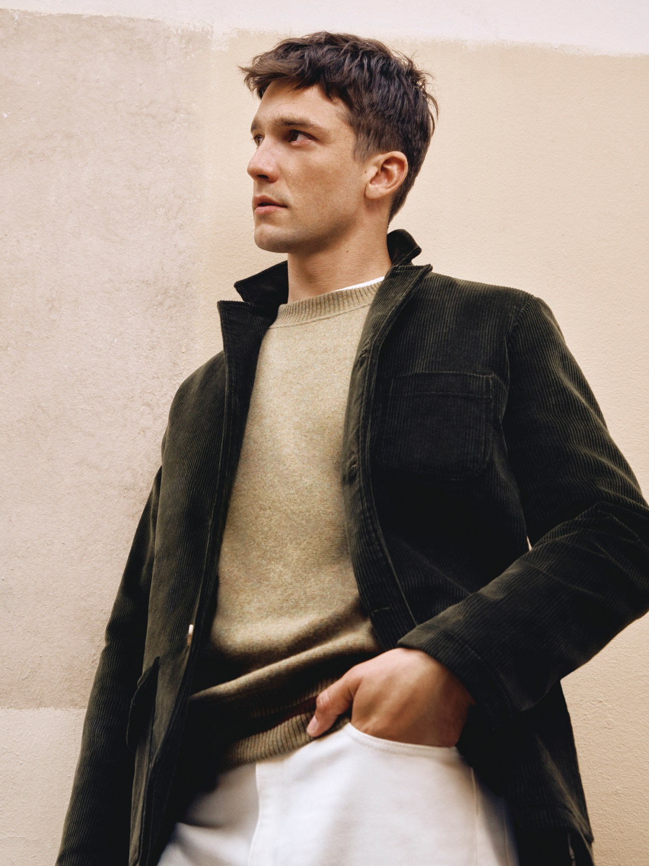 Model in brown corduroy jacket layered over cream jumper and white t-shirt with white trousers