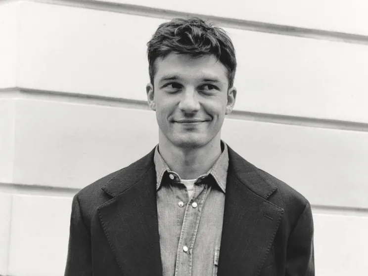 Model wearing dark blazer over light button-up shirt with hands in pockets against white paneled wall