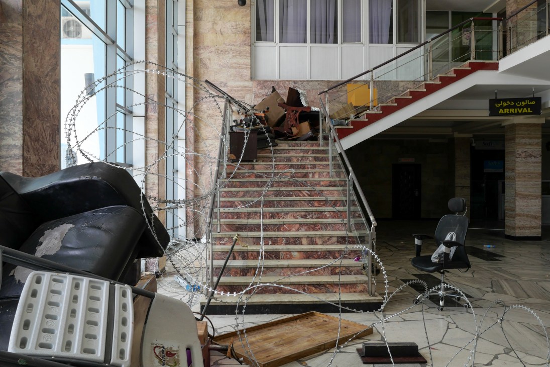 Interior view of damaged airport terminal with debris, overturned chairs, and razor wire scattered across the floor