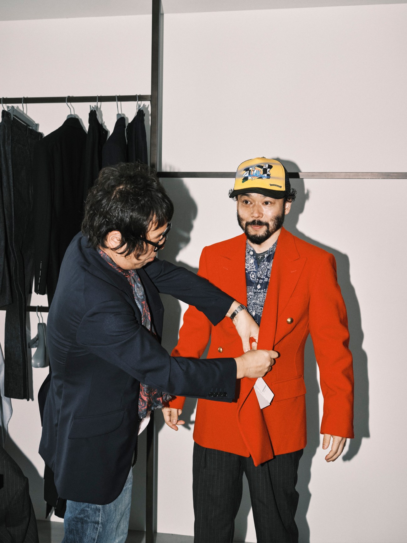 Two men in a clothing showroom, one wearing a dark navy blazer and the other wearing a bright orange blazer with a yellow and black cap, shaking hands or examining the orange jacket's fabric and construction.