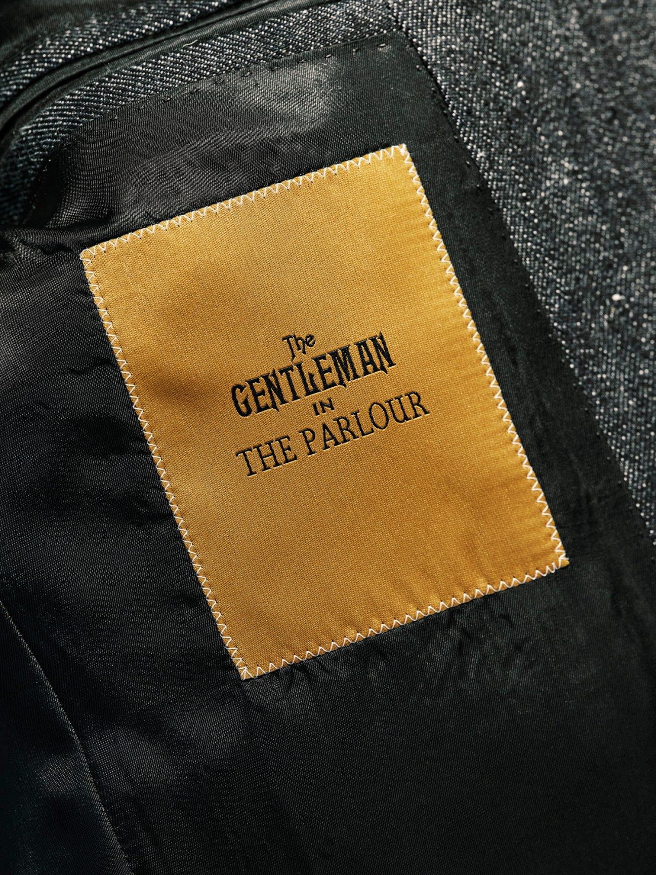 Close-up of a golden fabric label reading 'The GENTLEMAN in THE PARLOUR' sewn inside a dark jacket