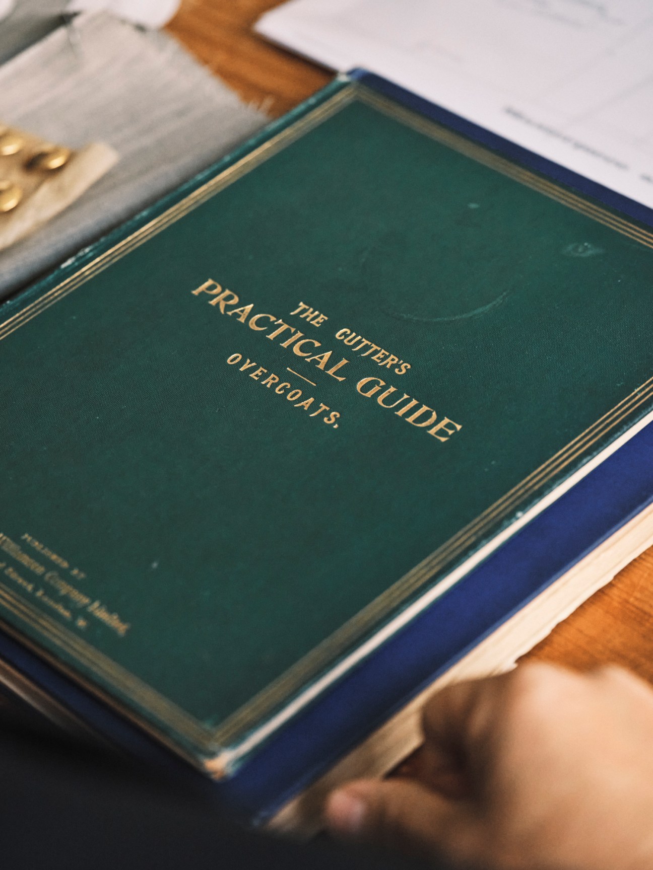 Open book with green cover titled 'The Cutter's Practical Guide to Overcoats' showing gold lettering on fabric