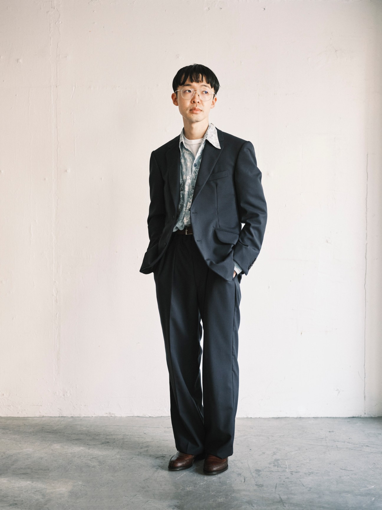Man in dark navy suit with white collar shirt standing against a plain white wall, hands in pockets, looking upward