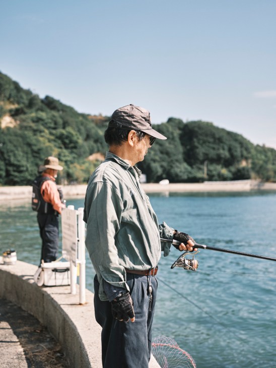 Man fishing from a dock overlooking water with forested hills in the background.