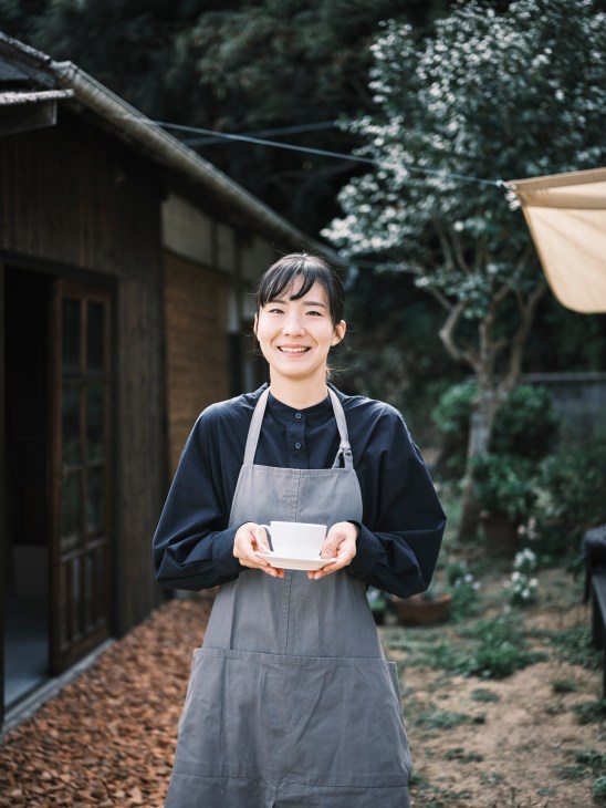 Young woman wearing a gray apron smiling while holding a white plate in a traditional Japanese courtyard.